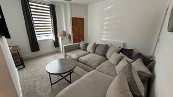 A living room with a sectional sofa, a round coffee table, a small wooden table, and a window with blinds.