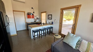 Een compacte keuken met een centraal eiland en barstoelen, een eethoek met een kleine tafel en stoelen, en een zithoek met een bank en een raam.