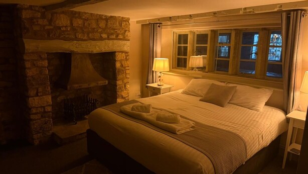 Un dormitorio acogedor con chimenea de piedra, una cama bien hecha con toallas, y una mesita de noche con lámpara.