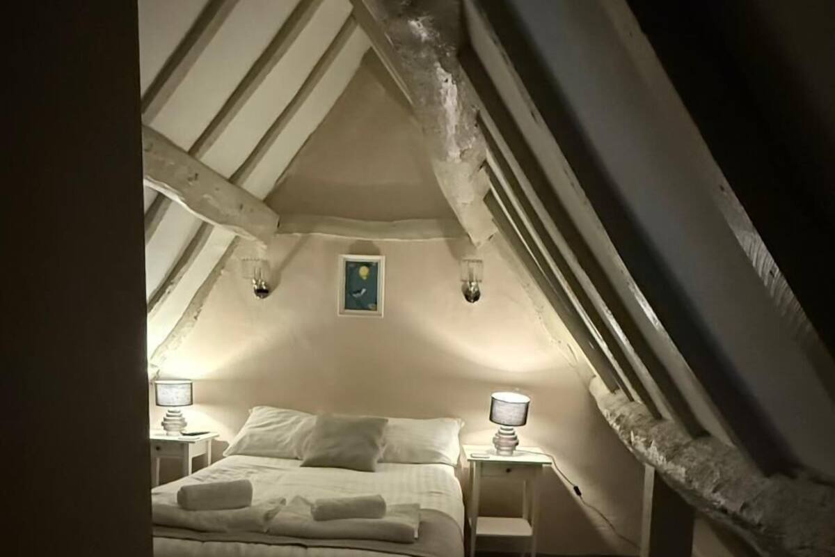 Um quarto de sótão aconchegante com teto inclinado, duas mesas de cabeceira com luminárias, uma cama com roupa de cama branca e uma pequena moldura na parede.