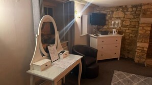 Una habitación con tocador, espejo, escritorio, silla, cómoda, televisor y paredes de piedra.