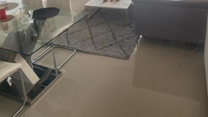 Un salon avec une table basse en verre, deux chaises blanches, un canapé gris et un téléviseur fixé au mur.