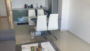 Un salon moderne avec une table basse en verre, un canapé gris et un coin cuisine à l’arrière-plan.