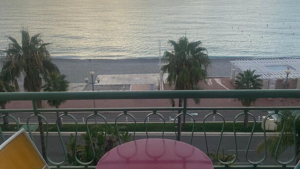 Un balcon avec deux chaises et une petite table, donnant sur une plage bordée de palmiers et sous un ciel dégagé.