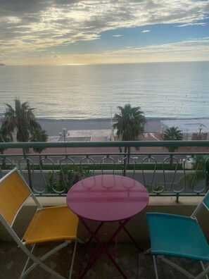Un balcone con due sedie e un tavolino, affacciato su una spiaggia con palme e cielo sereno.