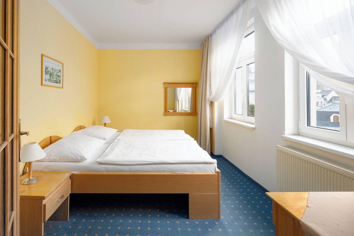A hotel room with a bed, bedside tables, a window with curtains, a radiator, and a picture on the wall.