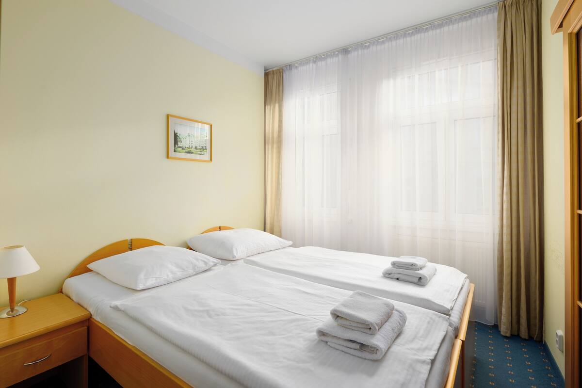 A hotel room with two beds, a bedside table with a lamp, and a large window with curtains.