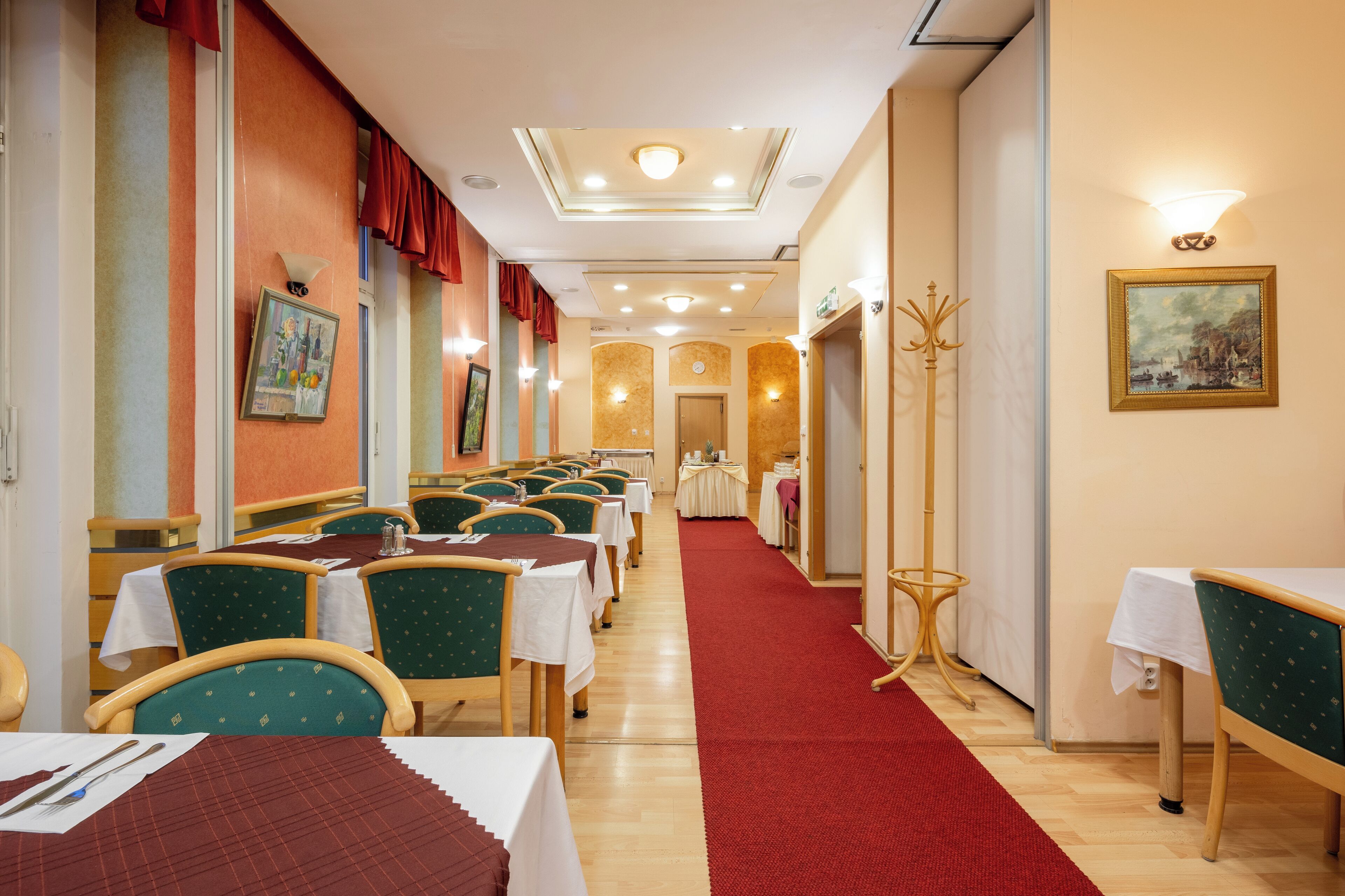 Uma sala de jantar de hotel com várias mesas e cadeiras, um tapete vermelho e obras de arte nas paredes.