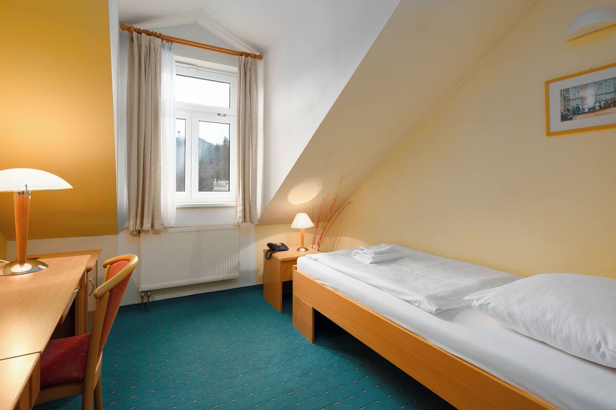 A small, well-lit room with a single bed, a desk, and a chair. There is a window with curtains, a lamp, and a framed picture on the wall.