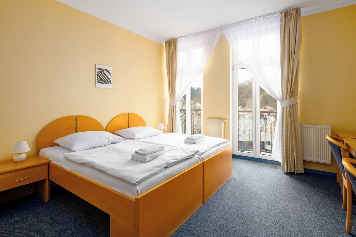 A hotel room with a large bed, two bedside tables with lamps, a view of the outside, and a small table with a chair.