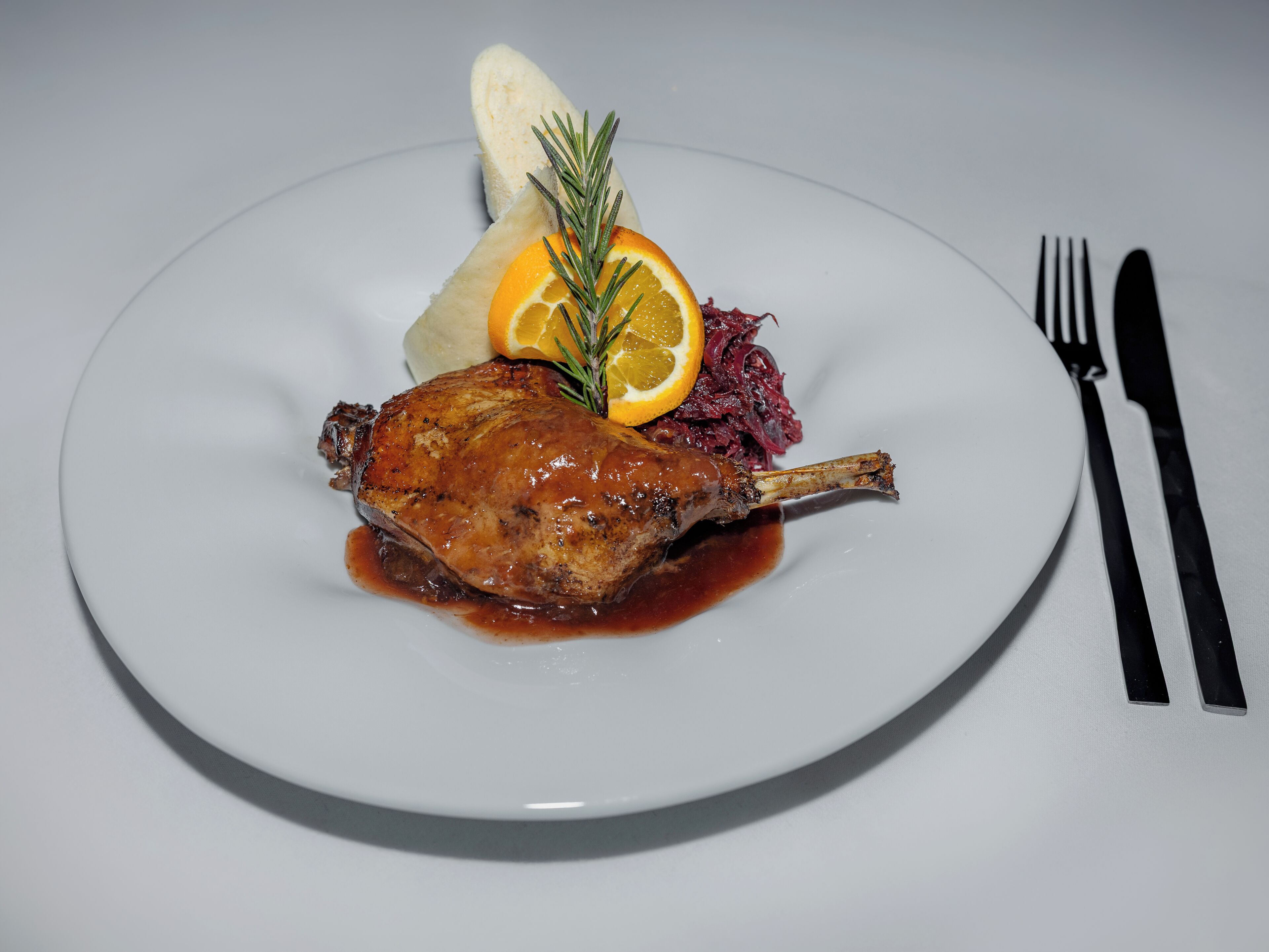Une assiette comprenant un morceau de cuisse de canard rôtie, une tranche d’orange et du chou rouge.