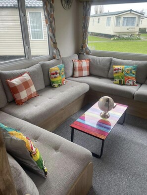 A living room with a grey sofa set, colorful pillows, a glass coffee table with a globe, and a view of a mobile home in the background. - Remarkable 1-bed Chalet in Minehead (Minehead)