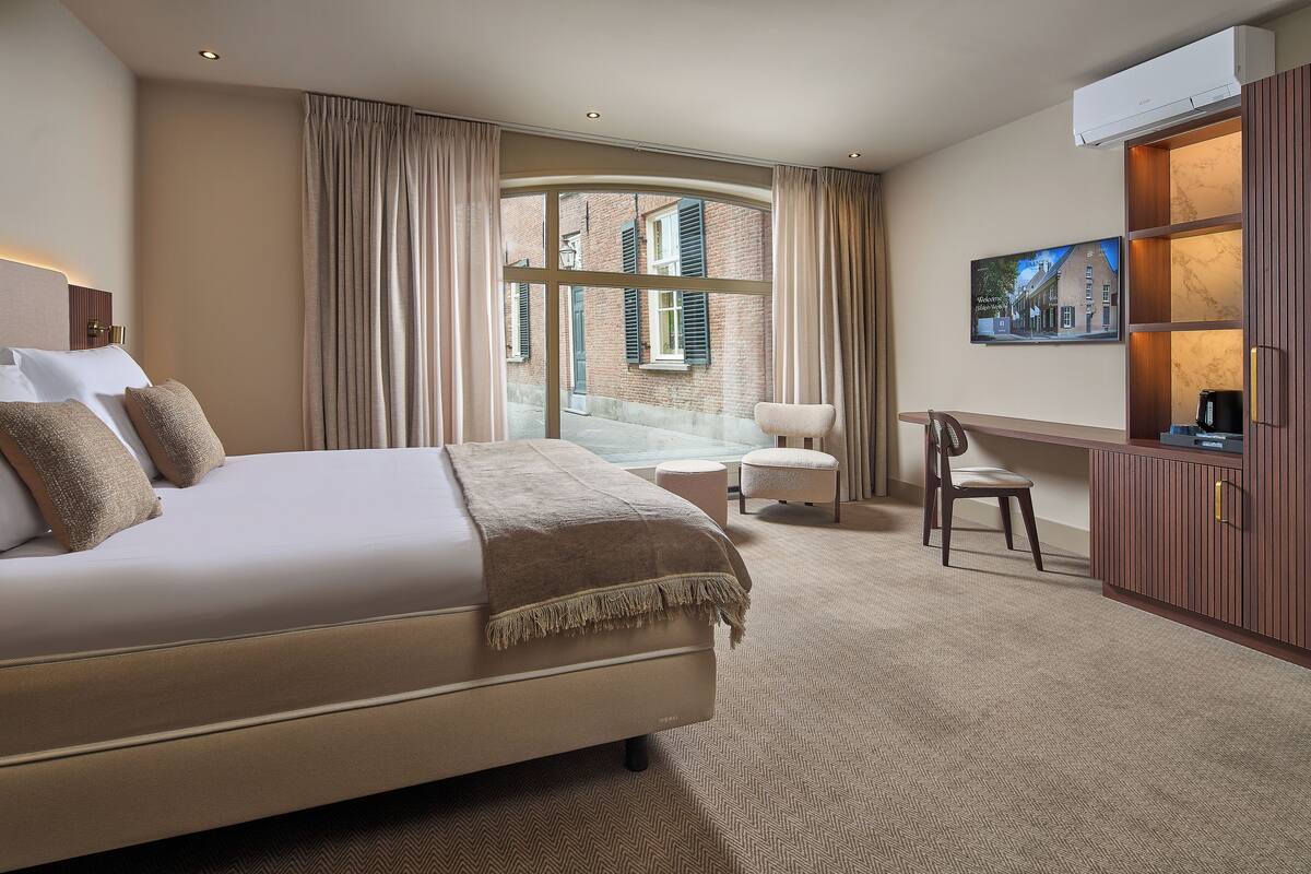 Habitación de hotel con una cama grande, una zona de estar con un sillón y reposapiés, un televisor y vistas a un edificio desde la ventana.