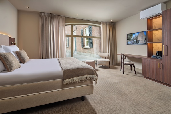 A hotel room with a large bed, a seating area with a chair and ottoman, a television, and a view of a building through the window. - The Saints Hotel ('s-Hertogenbosch)