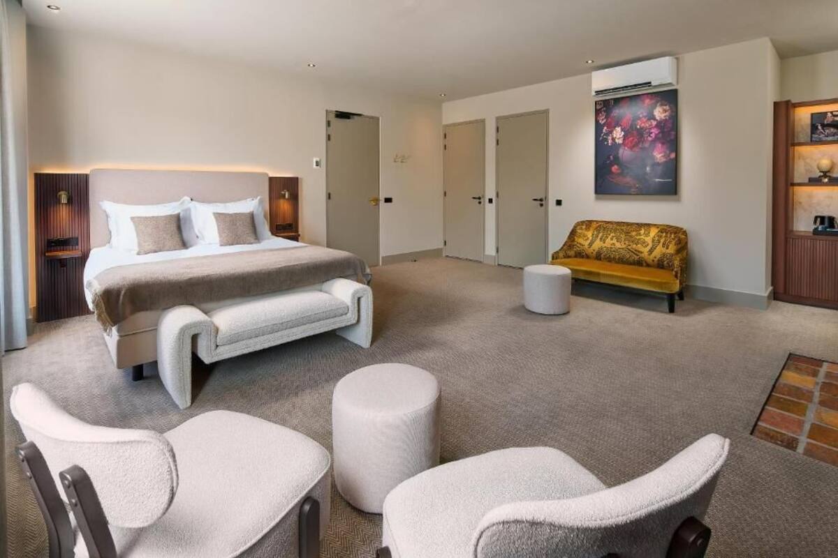 Habitación de hotel moderna con una cama grande, un sofá y dos sillones. Se observan varias puertas y un cuadro en la pared.