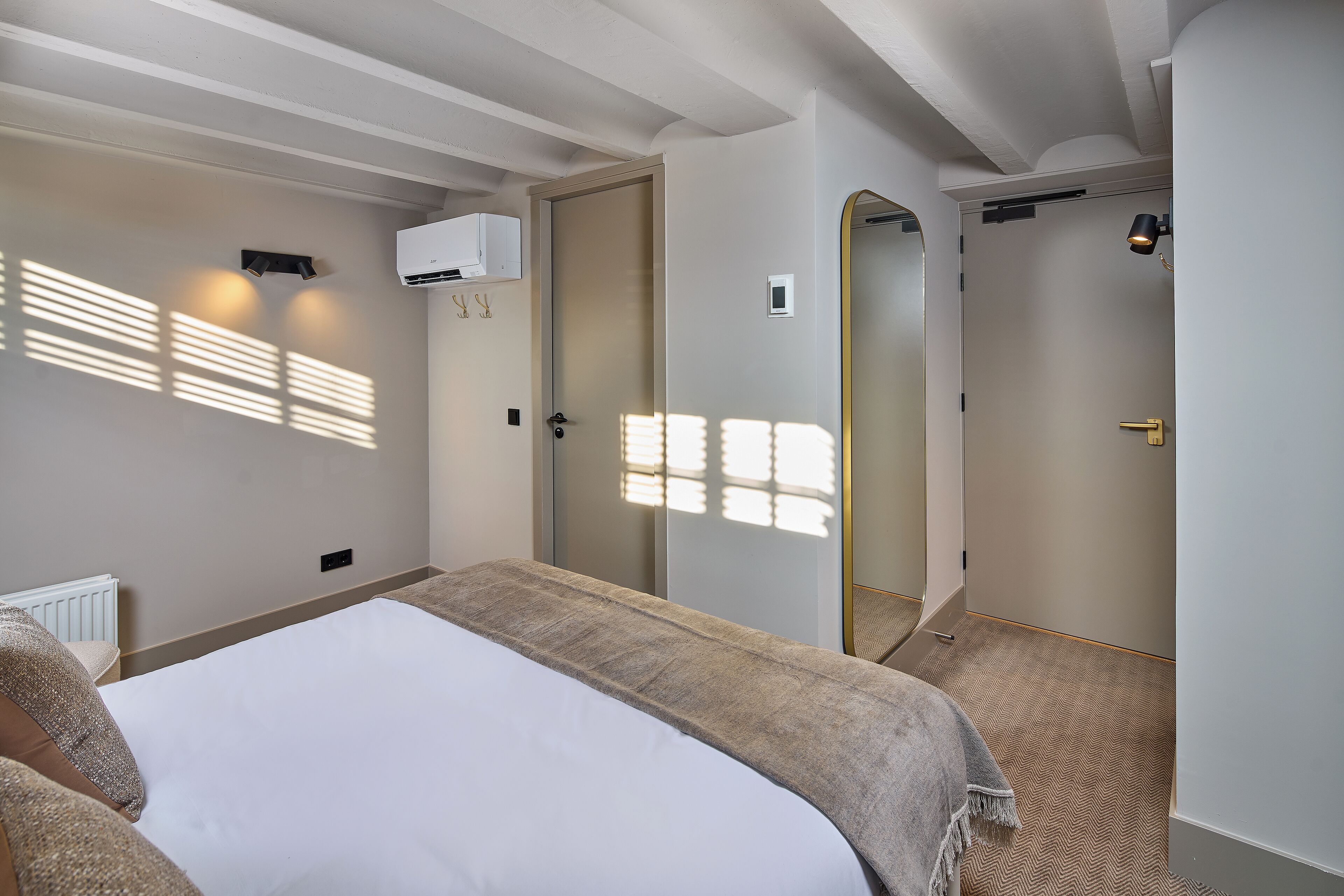 A hotel room with a bed, a radiator, an air conditioning unit, and a door leading to another room.