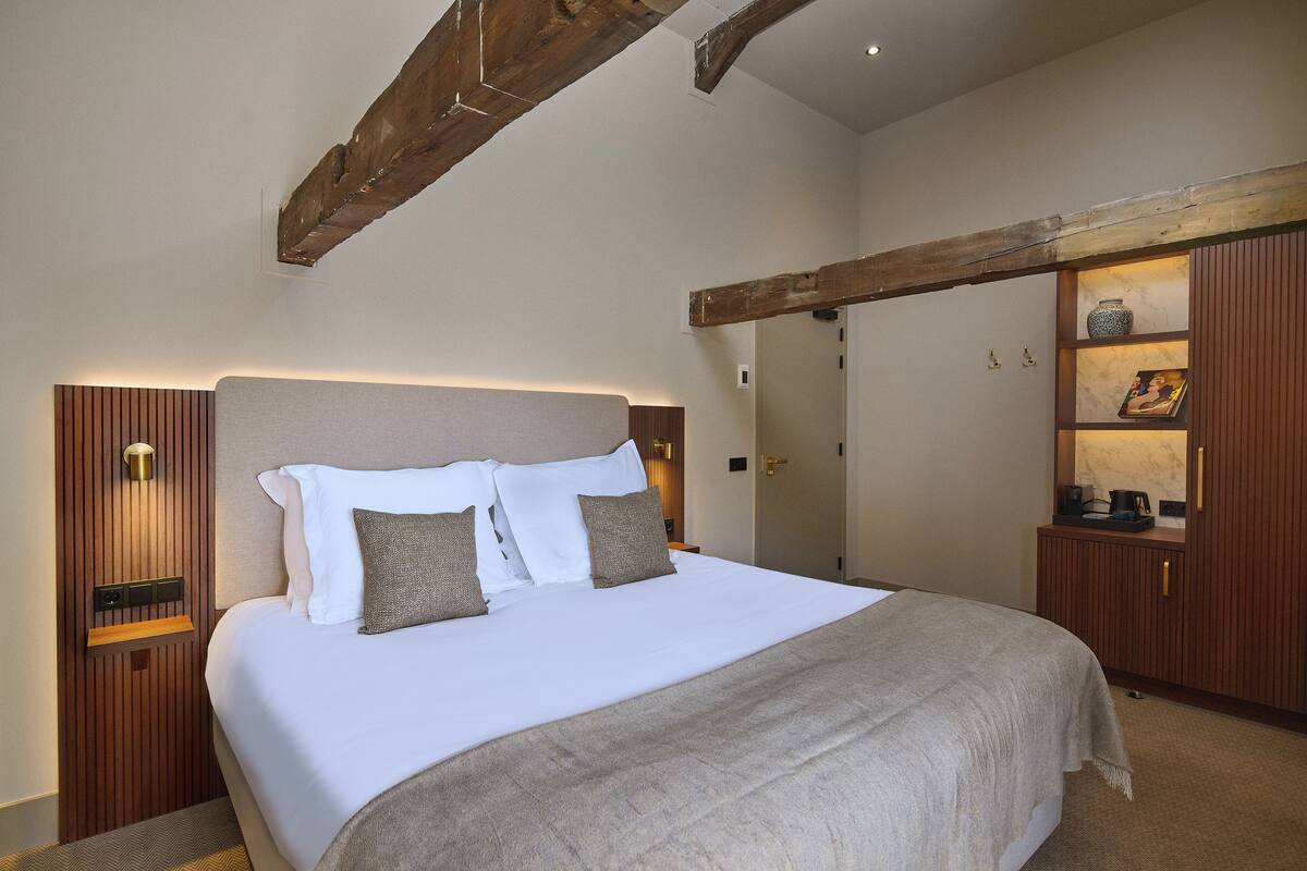 Una cama bien hecha con cabecera, una mesita de noche con lámpara y un armario de madera con espejo.