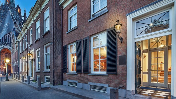 A row of brick buildings with large windows and shutters, a street lamp, and a building entrance with a glass door. - The Saints Hotel ('s-Hertogenbosch)