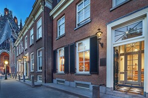 A row of brick buildings with large windows and shutters, a street lamp, and a building entrance with a glass door. - The Saints Hotel ('s-Hertogenbosch)