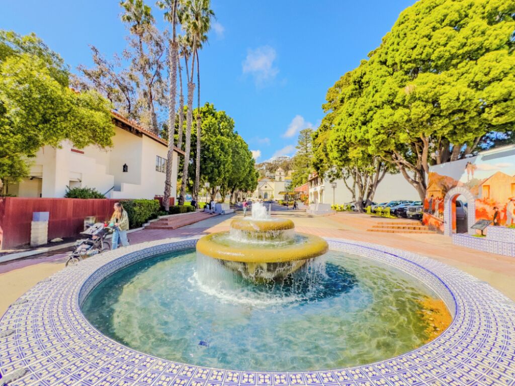 Una fuente en un patio con suelo de baldosas, rodeado de árboles y edificios.