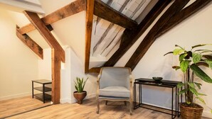 Ein Zimmer mit freiliegenden Holzbalken, einem hellgrauen Sessel, einem kleinen schwarzen Tisch, einer Topfpflanze und einem Korbwäscher.