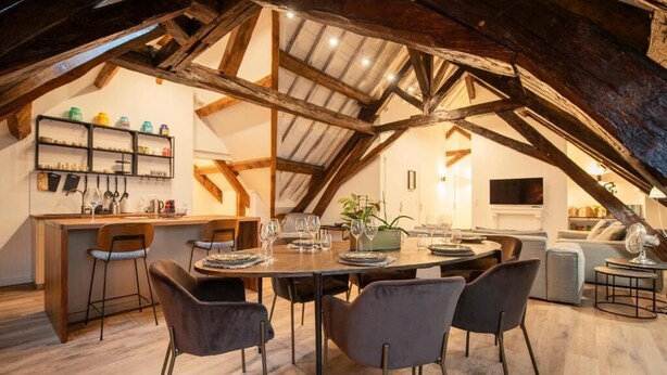 Ein Essbereich mit Holztisch und Stühlen, eine Bar mit Hockern und eine Küche im Hintergrund.