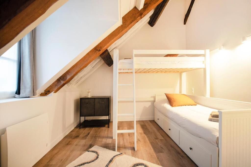 Un dormitori amb un llit superposat, una escala, una petita taula de nit i una finestra amb cortines.