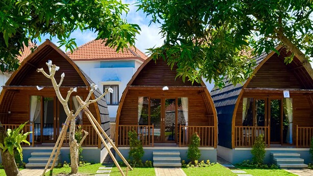 Una hilera de cabañas de madera con tejado de baldosas, un edificio central con fachada azul y un jardín cuidado con un árbol y una escalera de madera.