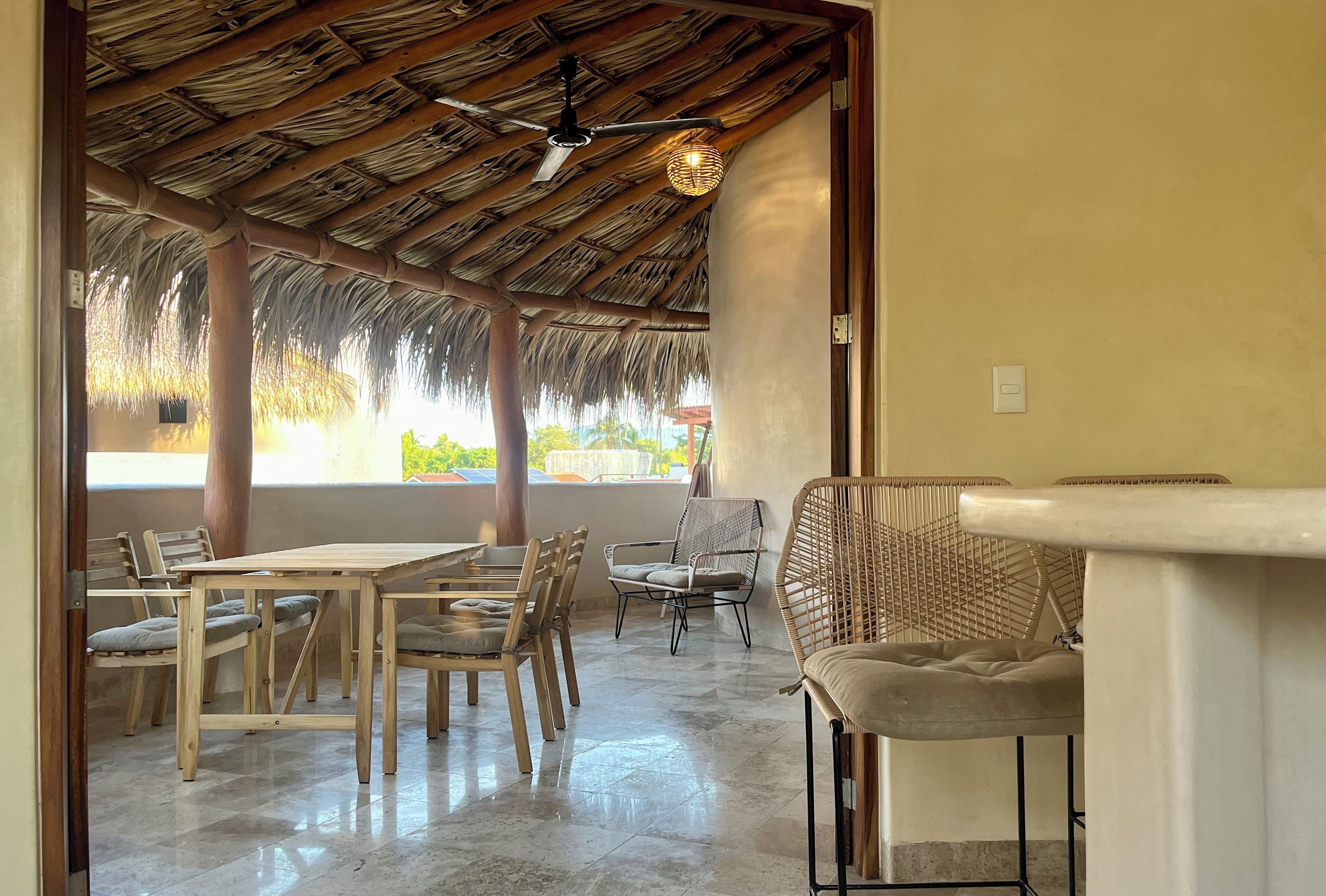 An indoor dining area with wooden tables and chairs, a thatched roof ceiling, and a view of the outdoors.