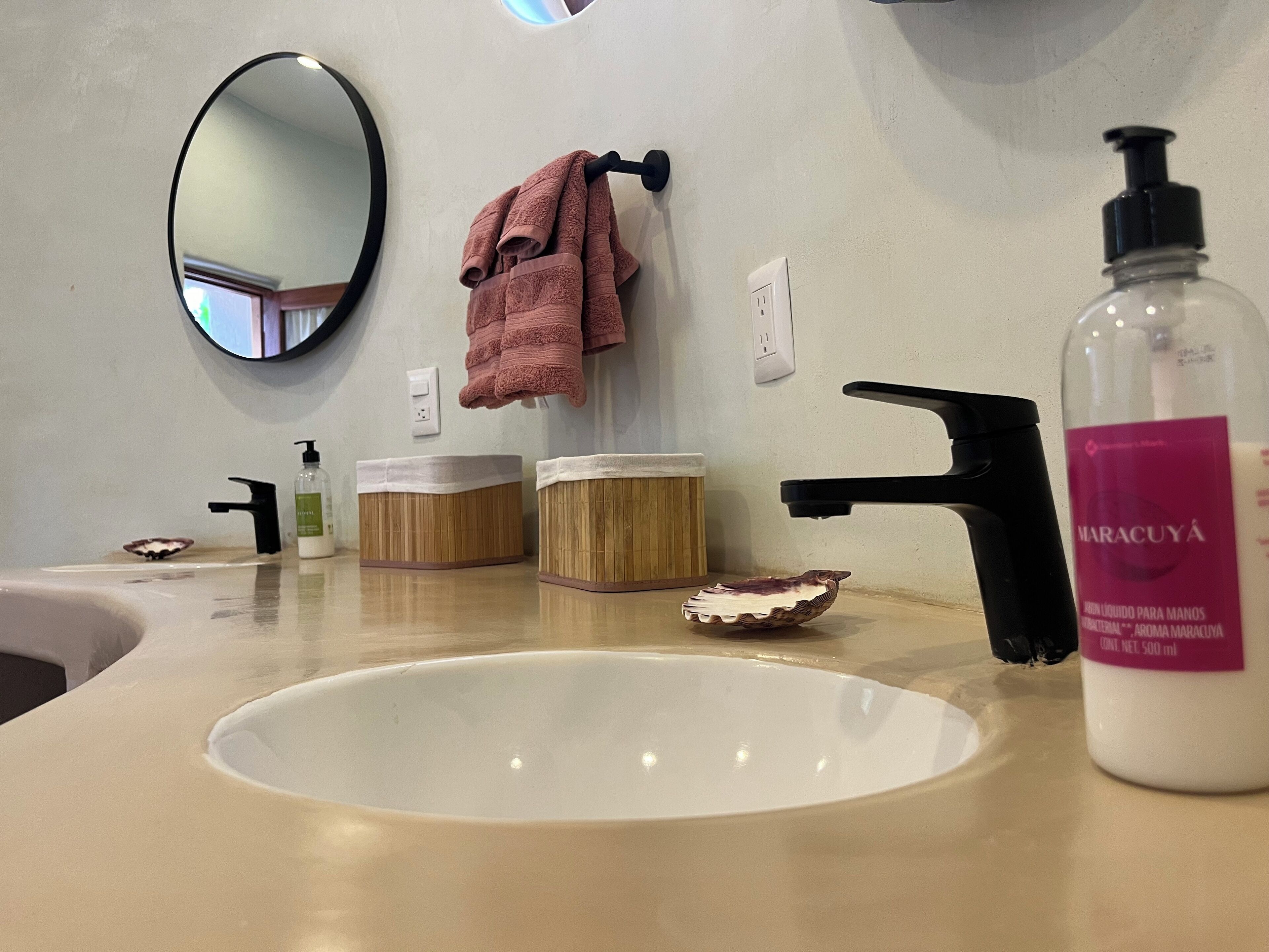 Un baño con un lavamanos blanco, un espejo, un perchero con una toalla rosa, dos recipientes de madera, un dispensador de jabón y una botella de jabón de manos Maracuya.