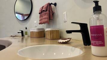 Un baño con un lavamanos blanco, un espejo, un perchero con una toalla rosa, dos recipientes de madera, un dispensador de jabón y una botella de jabón de manos Maracuya.