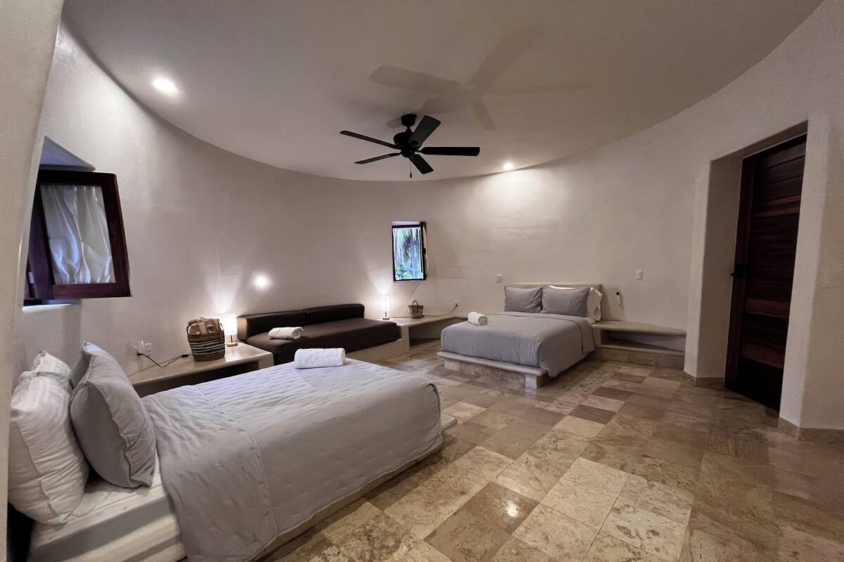 A modern hotel room with a curved ceiling, a ceiling fan, a large bed, a smaller bed, a seating area, and a television.