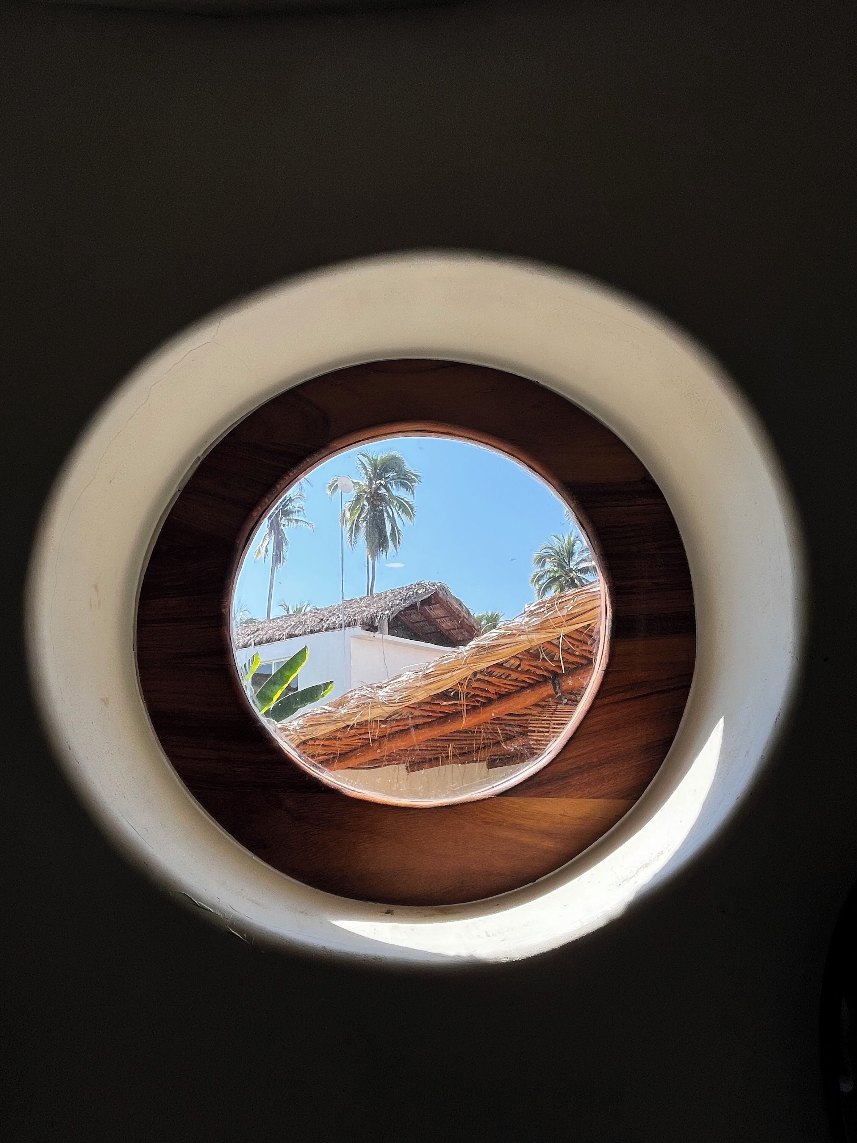 Una ventana circular con vistas a palmeras y una estructura con techo de paja.