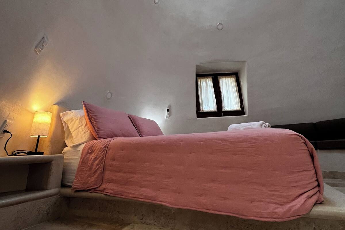 A bedroom with a bed, a nightstand, a lamp, a window, and a skylight.