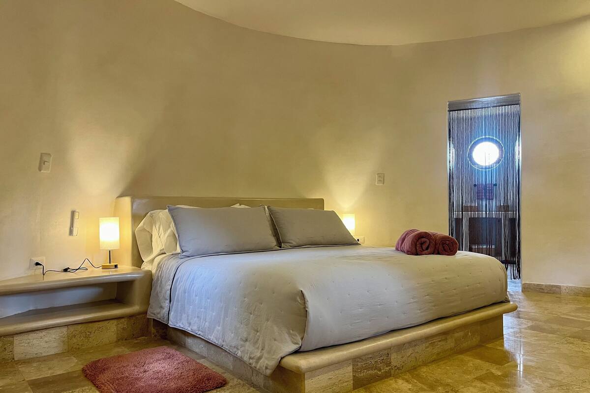 A neatly made bed with a headboard, a bedside lamp, and a red rug on a tiled floor.
