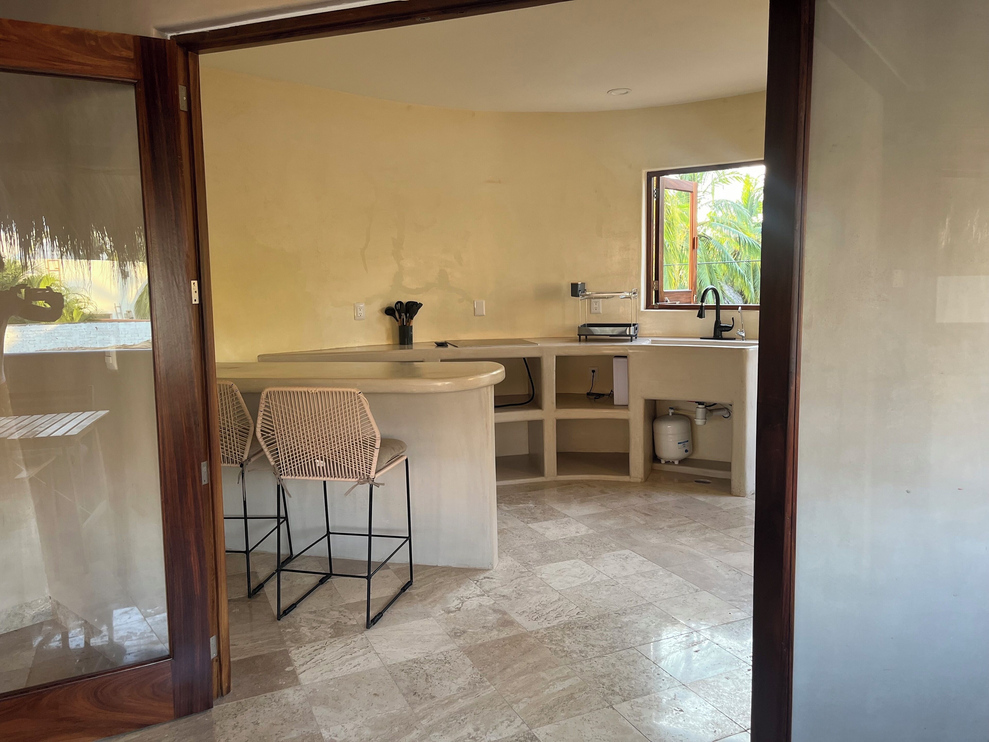 A kitchen with a white countertop, two wicker chairs, a window with a view of greenery, and a tiled floor.