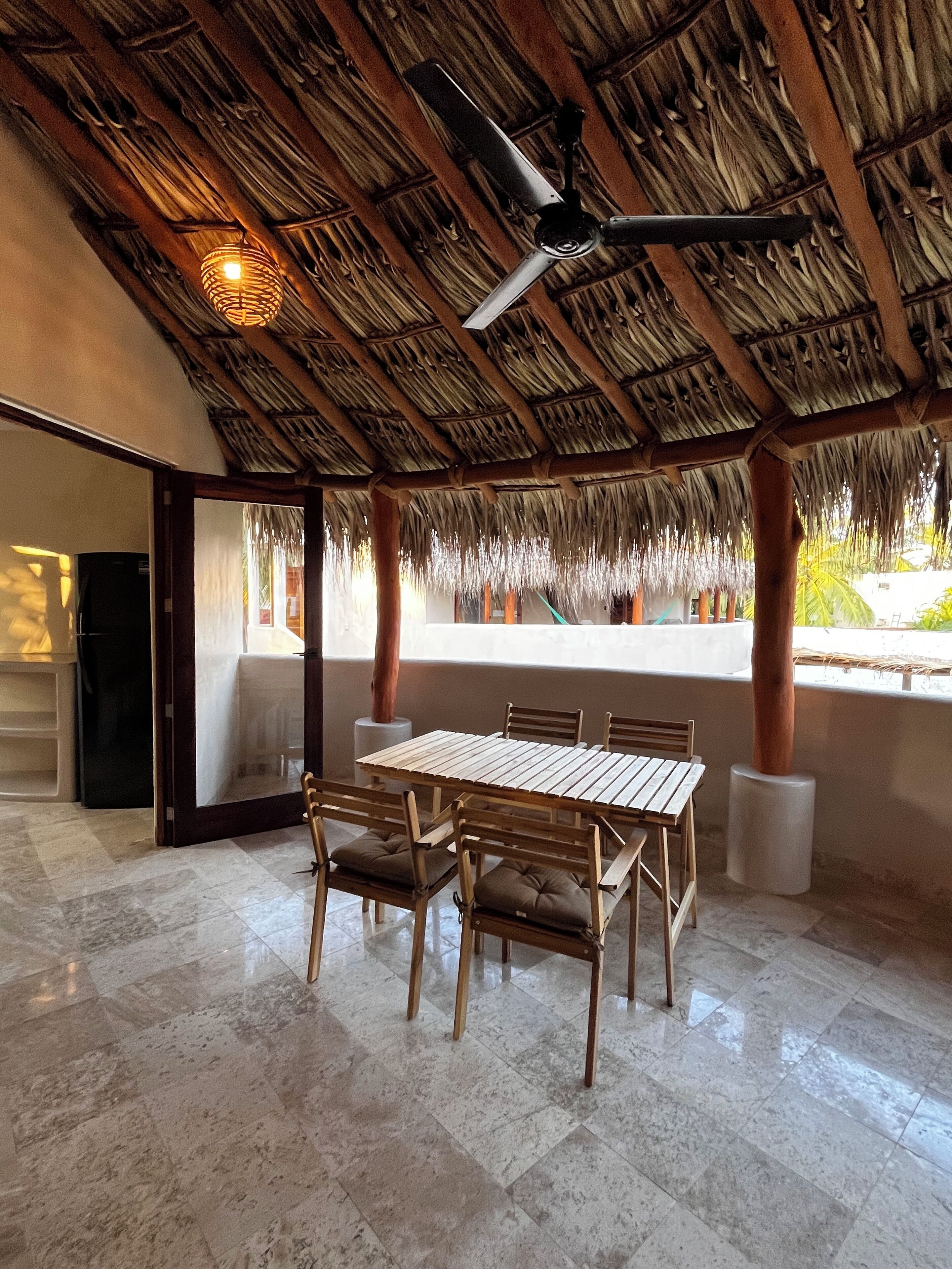 An outdoor patio with a thatched roof, a dining table with chairs, and a ceiling fan.