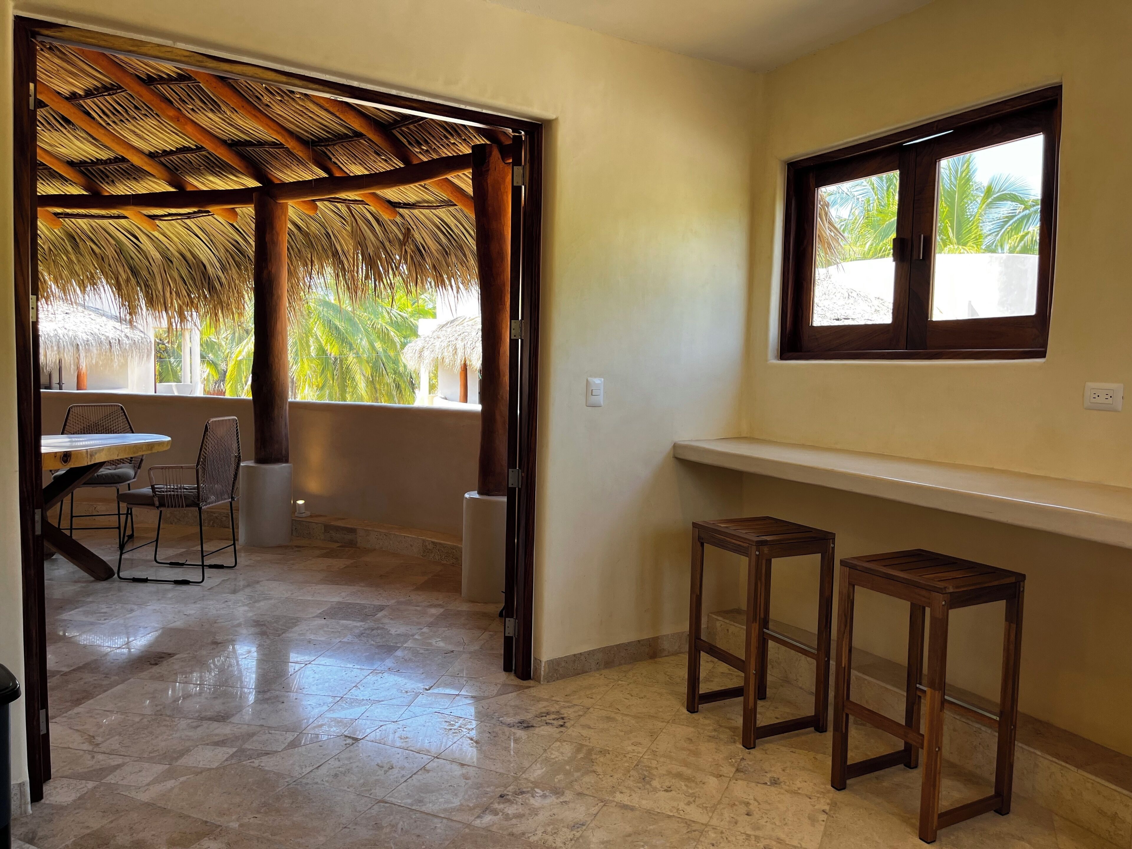 Una habitación con suelo de baldosas, estructura de techo de paja, ventana con vista a palmeras y un pequeño taburete de madera.