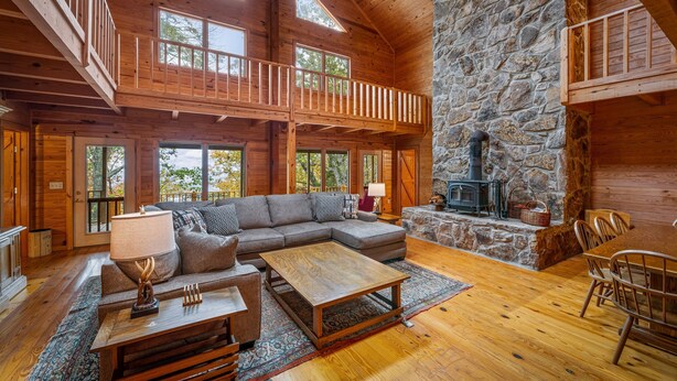 A spacious living room with a stone fireplace, wooden beams, and a large sofa. - Snail’s Pace South-6br/3.5 bath w/gorgeous views (Fort Payne)