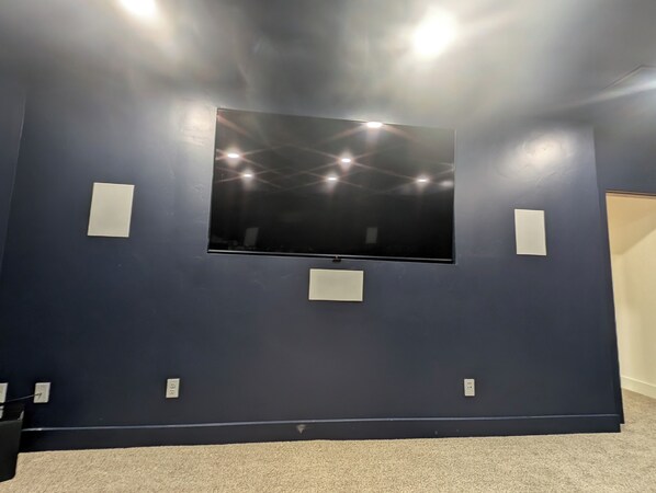 A room with a large flat-screen TV mounted on a dark blue wall, two small white panels, and carpeted flooring.