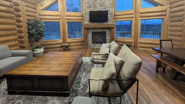 A log cabin interior with a stone fireplace, large windows, and a cozy seating area. - Remote Cabin near Warm River (Ashton)