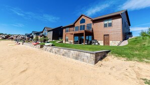 Uma casa moderna de dois andares com varanda, um muro de pedra e uma praia arenosa ao fundo.