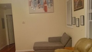 Een woonkamer met een grijze bank, een bruine leren bank en een houten salontafel. Er hangt een schilderij aan de muur en een deur naar een andere kamer.