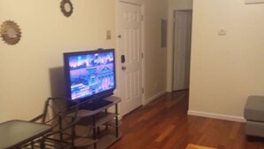 Een woonkamer met een televisie, een eettafel met stoelen, een salontafel, en een deur naar een andere kamer.