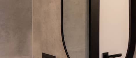 A modern bathroom with a large oval mirror, a black door handle, and a light switch on the wall.