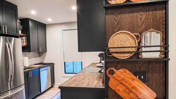 Una cocina moderna con armarios oscuros, un refrigerador de acero inoxidable, una encimera de madera y un estante empotrado de madera que exhibe diversos utensilios de cocina.