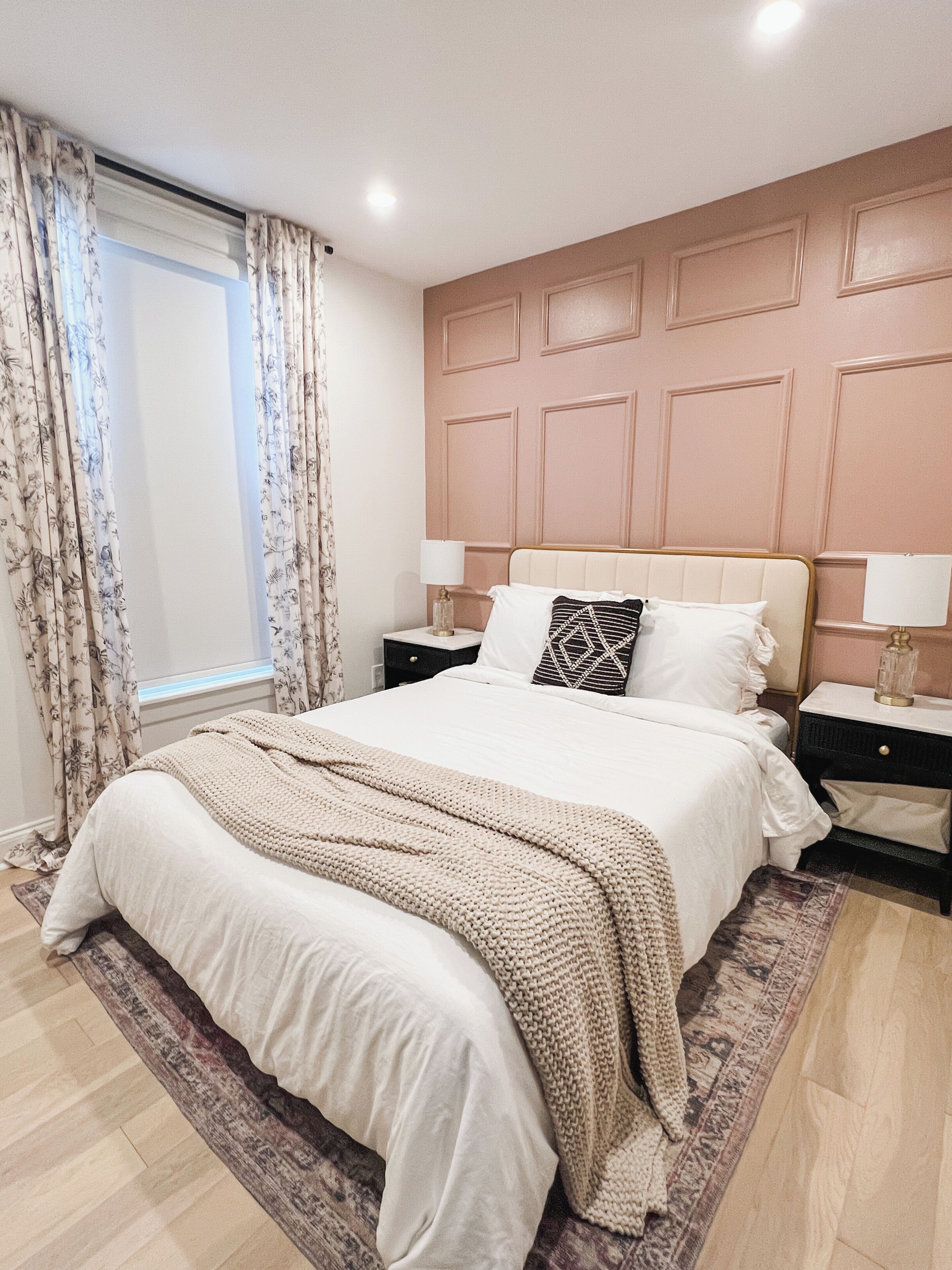 A bedroom with a large bed, two bedside tables, a window with curtains, and a headboard with paneling.
