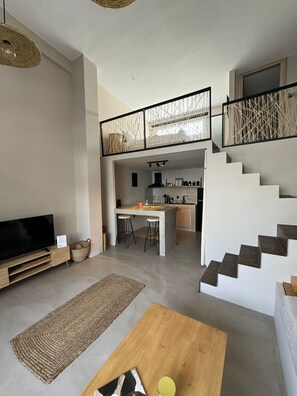 A modern living room with a TV, a coffee table, and a kitchen area with a dining table and stools. There is a staircase leading to an upper level.