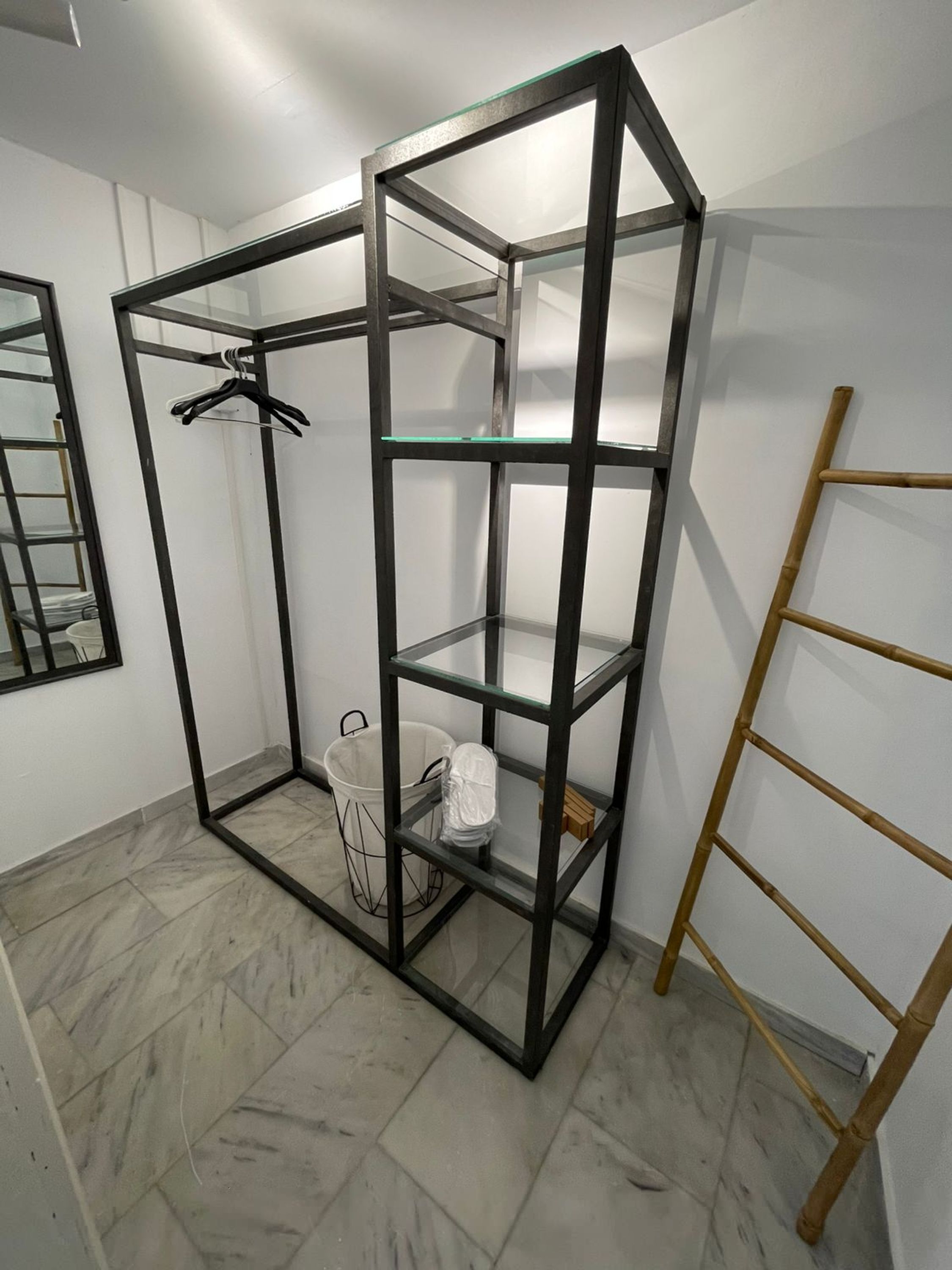 Una habitación con una estantería de metal, un cubo blanco y una escalera de madera.