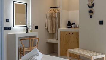 A hotel room with a bed, a wooden chair, a bench, a mirror, a sink, a wooden cabinet, and a wall-mounted towel rack.